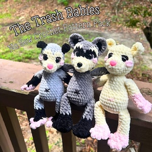 May include: Three plush crochet animal toys featuring the text The Trash Babies No Sew Crochet Pattern Pack. The left toy is light gray with black ears and feet, the center is dark gray with black markings, and the right is cream with pink feet.
