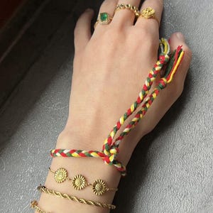 May include: A hand showcases multiple jewellery items. Gold rings with floral and gemstone designs are on the fingers. Several gold bracelets are visible, including a braided cord bracelet, a sunflower chain, and a gemstone bracelet. A braided cord bracelet in red, yellow, and green is also present.