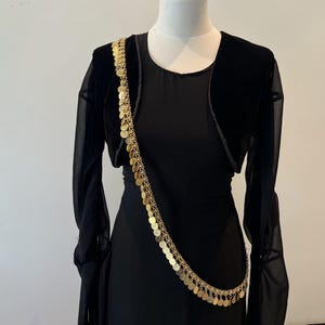 Könnte beinhalten: Ein schwarzes Kleid mit langen, transparenten Ärmeln und einem kurzen Bolero aus Samt, ergänzt durch eine lange, goldfarbene Kettenschärpe mit zahlreichen kleinen, runden, metallischen münzähnlichen Scheiben, die diagonal über den Oberkörper verläuft.