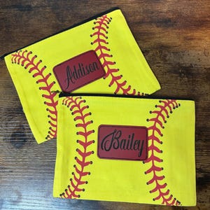 Könnte beinhalten: Zwei gelbe Softballetaschen mit Reißverschluss und roten Nahtdetails. Eine Tasche hat ein rotes Rechteck mit dem Namen "Addison" in schwarzer Schrift. Die andere hat "Bailey" in schwarzer Schrift.