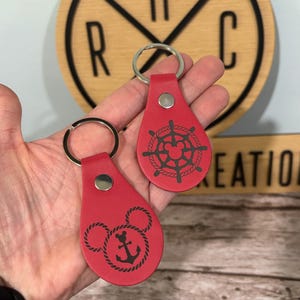 May include: Two red leather keyrings with silver rings. One keyring features a black anchor design with Mickey Mouse ears, and the other has a black ship's wheel design. The keyrings are held in a hand.