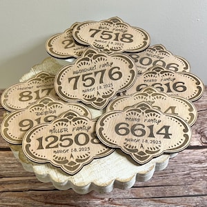May include: A close-up of a pile of wooden signs with engraved numbers and family names. The signs are decorated with intricate designs and the text "MARCH 1-8, 2025" is visible on several of the signs.