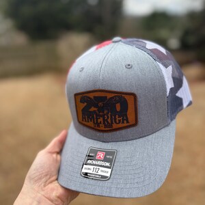 May include: A grey Richardson 112 trucker hat with a brown leather patch displaying an eagle and the text "20 AMERICA 1776-2020". The hat features a red, white, and blue camouflage pattern at the back and a curved peak. Adjustable.