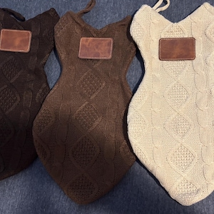 Puede incluir: Tres manoplas de horno de punto en color marrón oscuro, marrón medio y crema. Cada manopla tiene un patrón de diamantes y un parche de cuero marrón. Las manoplas tienen forma de vestido y tienen un lazo para colgar.