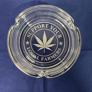 May include: Clear glass ashtray with a circular design. The center features a cannabis leaf and the text "SUPPORT YOUR LOCAL FARMERS" in white. The ashtray has three notches for resting items.