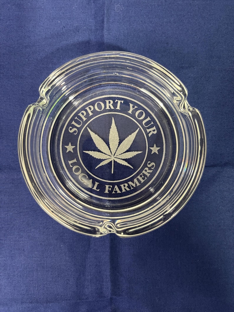 May include: Clear glass ashtray with a circular design. The center features a cannabis leaf and the text "SUPPORT YOUR LOCAL FARMERS" in white. The ashtray has three notches for resting items.