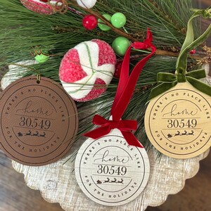 Può includere: Tre ornamenti natalizi rotondi con la scritta "THERE'S NO PLACE LIKE HOME 30549 FOR THE HOLIDAYS". Uno è in pelle marrone, uno in legno e uno in legno chiaro. Ognuno ha un nastro rosso o un nastro di velluto verde per appenderlo. Accenti decorativi natalizi sullo sfondo.