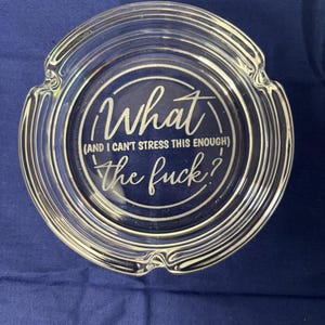 May include: Clear glass ashtray with a circular design. The text in the center reads "What (and I can't stress this enough) the fuck?". The ashtray has a ridged edge and is set against a blue background.