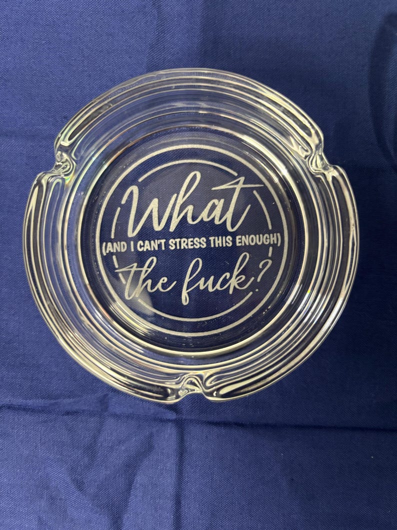 May include: Clear glass ashtray with a circular design. The text in the center reads "What (and I can't stress this enough) the fuck?". The ashtray has a ridged edge and is set against a blue background.