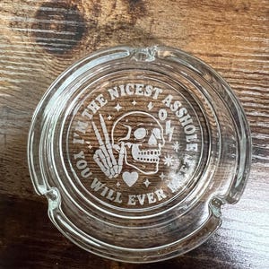 May include: Clear glass ashtray with a circular design. The ashtray features the text "I'M THE NICEST ASSHOLE YOU WILL EVER MEET" surrounding a skull, peace sign, heart, and lightning bolt.