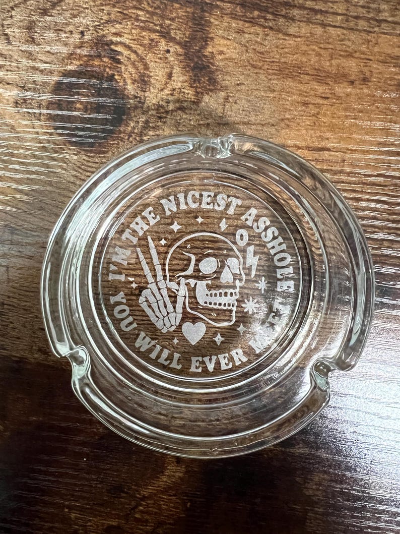 May include: Clear glass ashtray with a circular design. The ashtray features the text "I'M THE NICEST ASSHOLE YOU WILL EVER MEET" surrounding a skull, peace sign, heart, and lightning bolt.