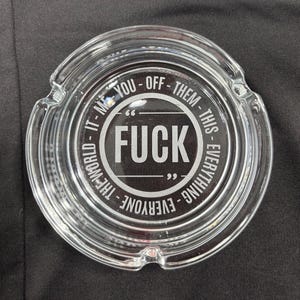 May include: Clear glass ashtray with a circular design. The center features the word "FUCK" in bold white letters, surrounded by the phrases "IT - ME - YOU - OFF - THEM - THIS - EVERYTHING - EVERYONE - THE WORLD".