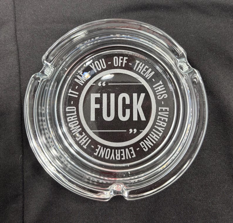 May include: Clear glass ashtray with a circular design. The center features the word "FUCK" in bold white letters, surrounded by the phrases "IT - ME - YOU - OFF - THEM - THIS - EVERYTHING - EVERYONE - THE WORLD".
