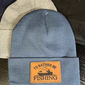 Peut inclure: Un bonnet en tricot bleu avec un patch en cuir marron qui dit "J'aimerais mieux être à la pêche" avec une silhouette de bateau et une personne à la pêche.
