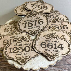 May include: A close-up of a pile of wooden plaques with engraved family names and dates. Each plaque features a unique design with a number and the text "MARCH 1-8, 2025".