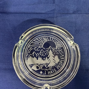 May include: Clear glass ashtray with a circular design featuring a mountain landscape, sun, and the text "I HATE PEOPLE". The ashtray has a rounded shape with three notches for resting cigarettes.