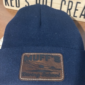 Op de afbeelding: Een marineblauwe gebreide muts met een bruine leren patch. De patch toont de tekst "MUFF'S Diving School" en een zwemmerafbeelding. Een winteraccessoire.