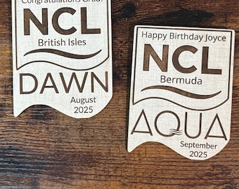 Könnte beinhalten: Zwei Holzschilder mit graviertem Text. Eines lautet "Herzlichen Glückwunsch zum Abschluss!" mit "NCL British Isles" und "DAWN August 2025." Das andere sagt "Alles Gute zum Geburtstag Joyce" mit "NCL Bermuda" und "AQUA September 2025."