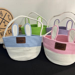 Op de afbeelding: Vier kleurrijke paasmanden met konijnenoren. De manden zijn blauw, groen, paars en roze, met witte en gekleurde secties. Elke mand heeft een handvat en een naamlabel. Een houten bord is zichtbaar op de achtergrond.