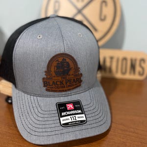 Puede incluir: Gorra trucker gris con parte trasera de malla negra y visera curva. La parte delantera presenta un parche de cuero marrón con un gráfico de barco y el texto "THE BLACK PEARL CRUISE LINES". Una etiqueta Richardson 112 Trucker es visible.