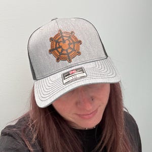 Puede incluir: Gorra de camionero gris con parte trasera de malla negra y un parche de cuero marrón con un diseño de rueda náutica con una silueta de Mickey Mouse. La gorra tiene una visera curva y una etiqueta blanca.