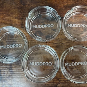 May include: Five clear glass ashtrays with the word "MUDDPRO" etched in the center. Each ashtray has a circular design with a small notch for resting cigarettes. The ashtrays are arranged on a dark wooden surface.