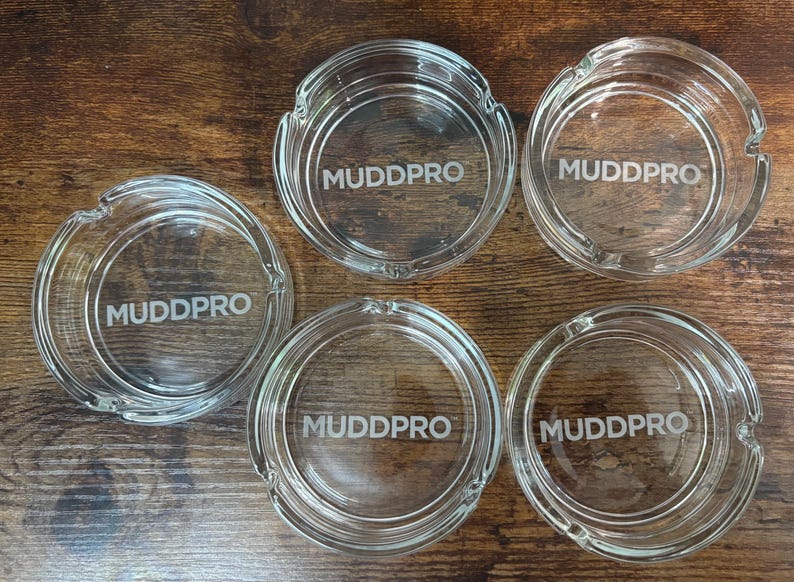 May include: Five clear glass ashtrays with the word "MUDDPRO" etched in the center. Each ashtray has a circular design with a small notch for resting cigarettes. The ashtrays are arranged on a dark wooden surface.
