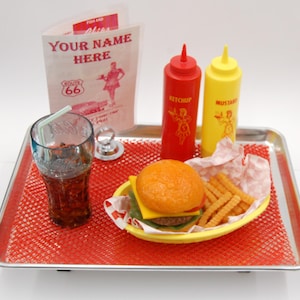 May include: A metal serving tray with a red mesh liner holding a cheeseburger, crinkle-cut fries in a yellow basket, a glass of dark soda with a straw, and red and yellow squeeze bottles labeled KETCHUP and MUSTARD, plus a Route 66 menu card.