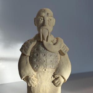 Puede incluir: Figura beige de un guerrero, de pie sobre una base rectangular. La figura tiene un largo bigote y barba, lleva armadura y una túnica. Las manos del guerrero están juntas frente a él.