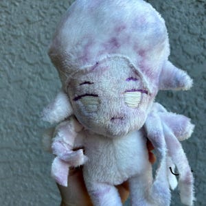 May include: A soft, plush doll in lavender and white hues, with a rounded head and multiple tentacle-like appendages. The doll has embroidered facial features, including closed eyes and a small mouth. The toy is made of a fuzzy material.