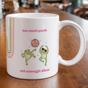 Può includere: Tazza in ceramica bianca con un design rosa e verde. La tazza presenta due rane che ballano con una palla da discoteca e il testo "too much panic not enough disco".