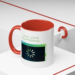 May include: White ceramic mug with a red interior and handle. The mug features the text "DAD JOHE BUFFERING..." and a graphic of a loading symbol on a dark blue background. The mug is sitting on a white surface.