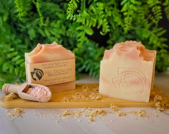 May include: Two bars of handmade soap with a pink and white swirl design, labeled "PIEL DESNUDA Makeup Remover." A wooden scoop holds pink powder, and the soap sits on a wooden board. The background features green foliage.