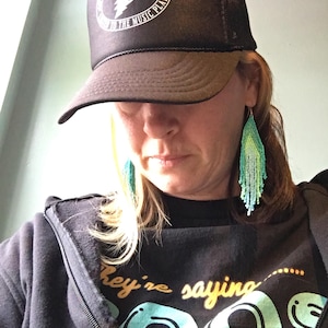May include: A black trucker hat with a white graphic and text that reads "DON'T GET CONFUSED LISTEN TO THE MUSIC PLAY". The person is wearing a black zip-up hoodie and beaded earrings. The hoodie has the text "They're saying" and a graphic.