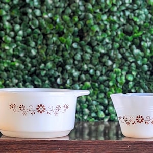 May include: Two white ceramic bowls with brown floral patterns. The larger bowl has handles and a wider base. The smaller bowl is tapered. Both are on a wooden surface against a green background.