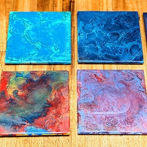 May include: Eight square coasters with abstract designs in shades of blue, teal, red, and purple. The coasters have a glossy finish and are arranged on a wooden surface. Each coaster features a unique, swirled pattern.