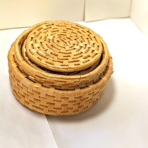May include: A set of three woven baskets in varying sizes. The baskets are made of light brown, natural-coloured woven material. The smallest basket serves as a lid for the medium-sized basket, which sits atop the largest basket.