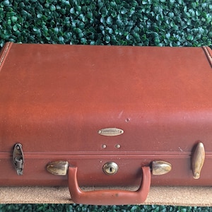 May include: A vintage brown Samsonite suitcase with brass fittings, including latches, a handle, and a keyhole. The suitcase is rectangular and shows signs of wear, indicating previous use.