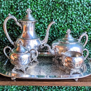 Puede incluir: Un juego de té plateado que incluye una tetera, una lechera, un azucarero y una bandeja. El juego se exhibe sobre una superficie de madera contra un fondo de follaje verde. La tetera tiene un asa curva y una tapa decorativa.