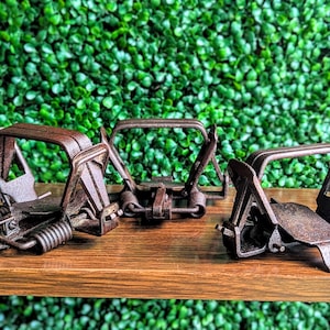 May include: Three vintage-style metal animal traps with a dark, rusted finish. Each trap features a spring-loaded mechanism, a flat trigger plate, and a hinged jaw frame. The traps are arranged in a row, showcasing their mechanical construction.