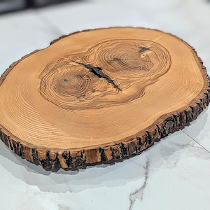 May include: A round, wooden serving board with a natural edge. The board is light brown with darker wood grain patterns and a dark crack in the center. The bark edge is rough and textured.