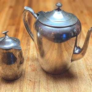 May include: A silver tea set, including a teapot and a creamer, sits on a wooden surface. The teapot has a curved spout and a lid with a decorative finial. The creamer has a handle and a matching lid. The set is reflective and appears vintage.