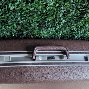 May include: A brown vintage suitcase with a textured surface, a brown handle, and silver accents. The suitcase has a white trim and a key lock. The overall design is classic and durable, suitable for travel.