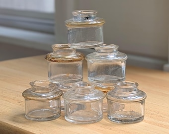May include: A collection of six small, clear glass inkwells arranged in a pyramid stack. Each vessel features a wide, flat rim and a cylindrical body with a slightly tapered neck. The glass is transparent with subtle variations in thickness and texture.