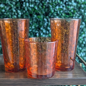 Puede incluir: Tres vasos para beber teñidos de naranja de diferentes tamaños. Los vasos tienen una superficie texturizada y estampada. Los vasos están dispuestos sobre una estantería de madera oscura frente a un fondo verde frondoso. Los vasos están vacíos.