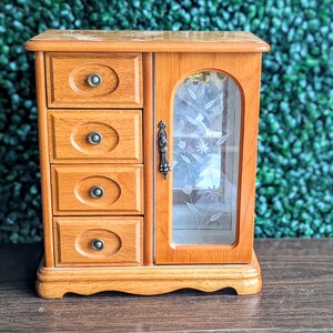 May include: A wooden jewelry armoire featuring four stacked drawers with oval recessed panels and small round knobs. To the right, a hinged door with a glass pane displays an etched floral design and a decorative metal handle for access.