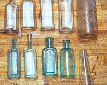 Puede incluir: Una colección de botellas de vidrio antiguas de varias formas y tamaños, dispuestas sobre una superficie de madera. Algunas botellas son transparentes, otras están teñidas de azul o marrón. Una botella tiene el texto "Allens Bottling Co.".