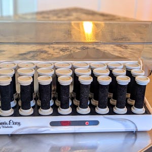 May include: A white and black hair setter case with a clear plastic lid, containing twenty black hair rollers with beige end caps. The front of the white base features the text "Free'n Easy by Windmere" and a small red indicator light.