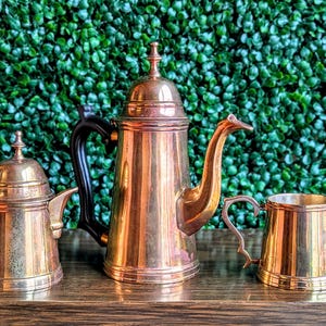 May include: A set of three antique brass tea or coffee service pieces. The set includes a small teapot, a tall coffee pot with a black handle, and a small creamer or sugar bowl. The pieces are displayed on a wooden surface against a green backdrop.