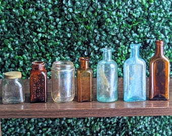 Puede incluir: Una colección de botellas y tarros de vidrio antiguos de varias formas y tamaños. Las botellas son de color marrón, ámbar y transparente, con una botella azul y un tarro con tapa de madera. Están dispuestas sobre una estantería de madera.